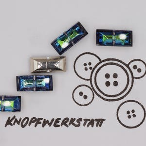 Op de afbeelding: Rechthoekige iriserende knopen en een zilveren knoop zijn gerangschikt op een wit oppervlak. Een zwarte illustratie van knopen staat naast de knopen. De tekst "KNOPFWERKSTATT" is zwart gedrukt.