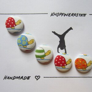 Könnte beinhalten: Fünf handgemachte Stoffknöpfe in Form von Schildkröten mit verschiedenen Mustern und Farben. Die Knöpfe sind in einer Reihe angeordnet, mit dem Text "KNOPFWERKSTATT" und "HANDMADE" sichtbar über und unter den Knöpfen.
