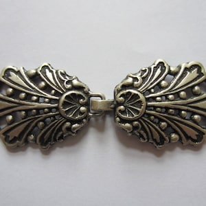 May include: A silver-toned metal clasp with a decorative design. The clasp is made of two pieces that connect in the middle. Each piece has a fan-shaped design with intricate details.