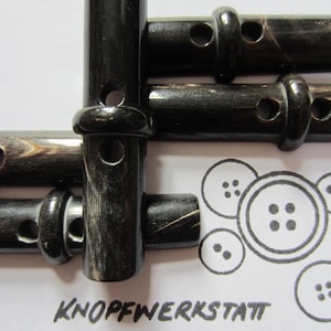 Könnte beinhalten: Vier schwarze und braune Hornknöpfe mit jeweils zwei Löchern. Die Knöpfe sind versetzt angeordnet. Die Knöpfe befinden sich auf einer weißen Oberfläche mit einer Zeichnung von vier Knöpfen.