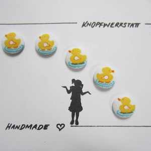 Può includere: Cinque bottoni di gomma gialla a forma di papera con acqua blu e becchi arancioni. I bottoni sono disposti a semicerchio attorno a una silhouette di una persona. Il testo "KNOPFWERKSTATT" e "HANDMADE" è visibile sullo sfondo bianco.