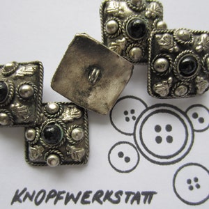 Könnte beinhalten: Vier silberfarbene quadratische Knöpfe mit schwarzen und grünen Akzenten. Die Knöpfe haben ein dekoratives Design mit einem seilartigen Rand und kleinen floralen Motiven. Die Knöpfe sind in einer Gruppe auf einer weißen Oberfläche angeordnet, unter der der Text "KNOPFWERKSTATT" steht.