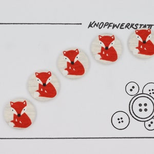 May include: Five white fabric buttons with red fox designs. The buttons are arranged in a diagonal line. The word "KNOFFWERKSTATT" is written in black at the top of the image.
