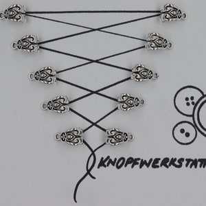 Puede incluir: Extremos de cordón de metal plateado con un diseño decorativo. Los extremos del cordón están dispuestos en un patrón de cruz sobre un fondo blanco. El texto "KNOFFWERKSTATT" está escrito en negro debajo de los extremos del cordón.