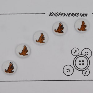 May include: Five white round buttons with brown kangaroo designs. The buttons are arranged in a semi-circle on a white background. The word "KNOFFWERKSTATT" is written in black at the top of the image.