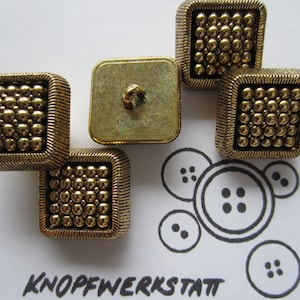 May include: Five gold-toned square buttons with a textured surface. The buttons have a raised, beaded design. The buttons are arranged in a cluster on a white surface.