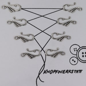 Puede incluir: Seis cierres de metal plateados con un diseño decorativo de espiral. Los cierres están dispuestos en dos filas de tres, con la fila superior ligeramente desplazada de la fila inferior. Los cierres están sobre un fondo blanco.