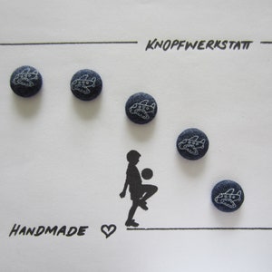 May include: Five blue denim buttons with white airplane designs. The buttons are arranged in a semi-circle around a silhouette of a person kicking a ball. The text "KNOPFWERKSTATT" and "HANDMADE" are written on the white background.