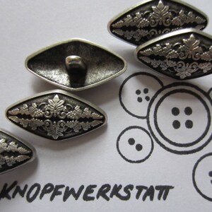 Könnte beinhalten: Fünf silberfarbene Knöpfe mit einem dekorativen floralen Design. Die Knöpfe sind diamantförmig mit abgerundeten Kanten. Die Knöpfe sind in einem Halbkreis auf einer weißen Oberfläche angeordnet. Der Text "Knopfwerkstatt" ist in Schwarz am unteren Rand des Bildes geschrieben.