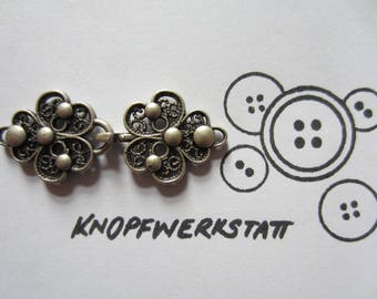 May include: Two silver filigree flower-shaped clasps with a connecting bar. The clasps are ornate and detailed, with a delicate, openwork design. The image also includes the text "Knopfwerkstatt".
