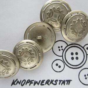 Op de afbeelding: Vier zilveren metalen knopen met een decoratief ontwerp. De knopen hebben een reliëf, ingewikkeld patroon van vier gestileerde bladeren in een ruitvorm. De knopen zijn in een halve cirkel op een wit oppervlak gerangschikt.