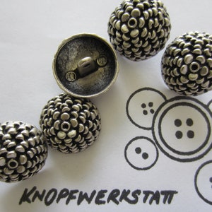 Op de afbeelding: Vijf zilverkleurige knopen met een getextureerd, verhoogd ontwerp. De knopen zijn rond en hebben een gat in het midden.