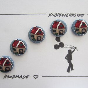 May include: Five handmade blue and red buttons with a gingerbread house design. The buttons are arranged in a row on a white card with the text "Knopfwerkstatt" and "Handmade" written in black.