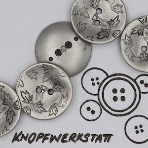 Puede incluir: Cinco botones de metal plateado con un diseño de hojas. Los botones están dispuestos en semicírculo. El texto "KNOFFWERKSTATT" está escrito en negro debajo de los botones.