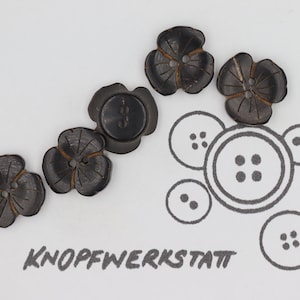 May include: Five black wooden buttons in the shape of a flower with a four-hole center. The buttons are arranged in a line on a white surface. The word "KNOFFWERKSTATT" is written in black below the buttons.