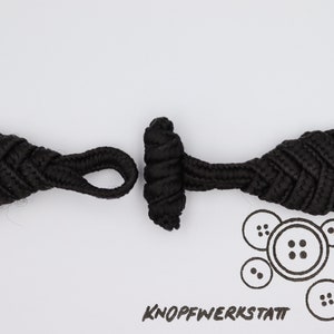 Pode incluir: Um par de fechos de botão de tecido preto. Os botões são feitos de um cordão trançado e têm um design de nó. Os botões são projetados para serem usados como fecho para roupas ou outros itens.