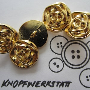 Puede incluir: Cuatro botones de metal de color dorado con un diseño de rosa. Los botones están dispuestos en semicírculo sobre una superficie blanca. La palabra "Knopfwerkstatt" está impresa en negro sobre la superficie.
