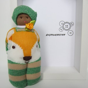 May include: A handmade sock doll with a green hat, orange and white striped clothing, and a fox face. The doll is standing on a white surface with a white frame behind it. The frame has the text "Knopfwerkstatt" and a design of buttons.