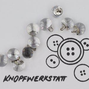 Può includere: Primo piano di un gruppo di bottoni in plastica trasparente con steli in metallo argentato. I bottoni sono disposti in una linea curva su una superficie bianca. La parola "KNOFFWERKSTATT" è scritta in inchiostro nero sotto i bottoni.