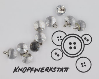 Könnte beinhalten: Nahaufnahme einer Gruppe von durchsichtigen Plastikknöpfen mit silbernen Metallstielen. Die Knöpfe sind in einer geschwungenen Linie auf einer weißen Oberfläche angeordnet. Das Wort "KNOFFWERKSTATT" ist in schwarzer Tinte unter den Knöpfen geschrieben.