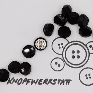Puede incluir: Botones negros facetados con un acabado brillante dispersos sobre una superficie blanca. También hay un botón con un centro plateado. La palabra "KNOPFWERKSTATT" está escrita en un estilo manuscrito. También se incluye un dibujo de botones.