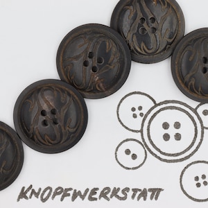 May include: Five dark brown wooden buttons with a carved floral design. The buttons have four holes each. The buttons are arranged in a row on a white surface. The word "KNOFFWERKSTATT" is written in black ink below the buttons.