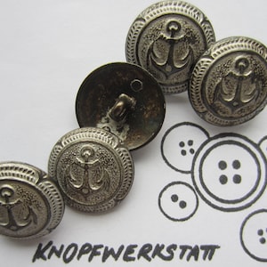 Peut inclure: Ensemble de boutons métalliques argentés avec un motif d'ancre. Les boutons ont un bord texturé et une boucle au dos pour la couture. L'image comprend également un croquis de modèles de boutons et le texte "KNOPFWERKSTATT".