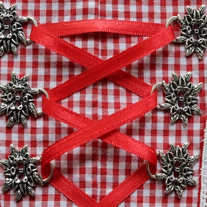 Könnte beinhalten: Rotes und weißes kariertes Stoff mit silbernen, blütenförmigen Metallknöpfen und roten Schleifenbändern.