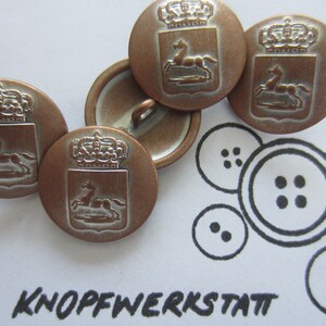 Puede incluir: Cuatro botones antiguos de color cobre con un diseño plateado. Cada botón presenta una corona, un escudo y un caballo. Los botones están dispuestos en semicírculo sobre una superficie blanca. El texto "KNOPFWERKSTATT" está escrito en tinta negra debajo de los botones.