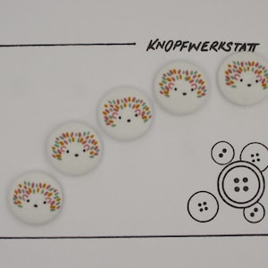 May include: Five white fabric buttons with a hedgehog design. The hedgehogs are pink, green, and yellow. The buttons are arranged in a line on a white background. The text "KNOFFWERKSTATT" is written in black ink at the top of the image.