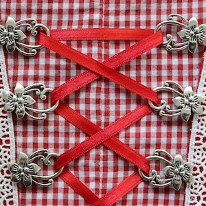Op de afbeelding: Rood en wit gingham-stof met witte kantversiering en zilveren metalen decoratieve sluitingen met bloemmotieven. Rood satijnen lint is door de sluitingen geregen.