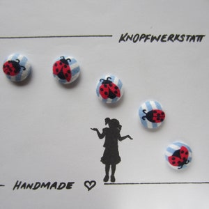Könnte beinhalten: Sechs Marienkäfer-Knöpfe mit blau-weißen Streifen. Die Knöpfe sind in einem Halbkreis um eine Personensilhouette angeordnet. Der Text "KNOPFWERKSTATT" und "HANDMADE" steht auf der Karte.
