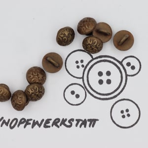 May include: A group of 9 gold-toned metal buttons with a decorative design. The buttons have a shank on the back. The buttons are arranged in a semi-circle on a white surface. The text "KNOPFWERKSTATT" is written in black ink below the buttons.