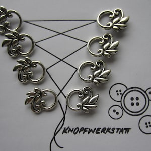 Puede incluir: Ocho adornos decorativos de metal plateado con diseño floral. Cada adorno tiene un lazo para sujetarlo a un proyecto. Los adornos están dispuestos en un patrón sobre una superficie blanca. El texto "KNOFFWERKSTATT" está escrito en tinta negra sobre la superficie.
