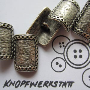 May include: Four silver metal buttons with a textured surface and a decorative border. The buttons are arranged in a semi-circle on a white surface with a black and white button design.