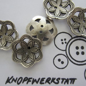May include: Five silver metal buttons with a floral design. The buttons have a cutout design and are in the shape of a flower.