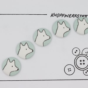 May include: Five light green fabric buttons with white fox faces on them. The buttons are arranged in a line, with the text "KNOFFWERKSTATT" above them.