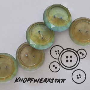 Könnte beinhalten: Fünf irisierende grün-gelbe Knöpfe mit jeweils zwei Löchern. Die Knöpfe sind in einem Halbkreis auf einer weißen Oberfläche angeordnet. Das Wort "KNOFFWERKSTATT" ist in Schwarz unter den Knöpfen geschrieben.