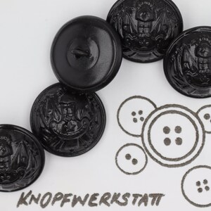 Può includere: Cinque bottoni neri con un designo ad aquila e un bottone nero semplice. I bottoni sono disposti a semicerchio su una superficie bianca. Il testo "KNOFFWERKSTATT" è scritto in inchiostro nero sotto i bottoni.