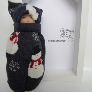 May include: A handmade doll crafted from a dark gray sock, featuring a snowman and snowflake design. The doll has a matching hat and scarf, with a visible face. The doll is displayed in a white frame, with the text "KNOPFWERKSTATT".