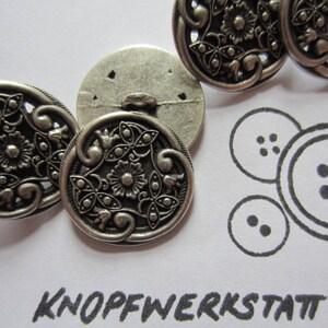 Könnte beinhalten: Vier silberne Metallknöpfe mit einem filigranen Blumendesign. Die Knöpfe haben eine erhabene, strukturierte Oberfläche und eine runde Form.