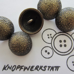 Puede incluir: Un conjunto de seis botones de color dorado antiguo con una superficie texturizada. Los botones son redondos y tienen un centro elevado. Los botones están dispuestos en círculo sobre un fondo blanco. La palabra "KNOPFWERKSTATT" está escrita en tinta negra debajo de los botones.