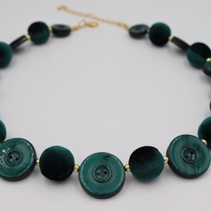 Può includere: Una collana di bottoni in velluto verde scuro e oro. La collana è composta da bottoni grandi e rotondi con un bordo dorato.
