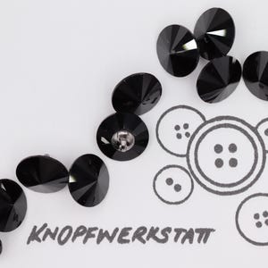 Puede incluir: Botones negros facetados con superficie reflectante dispersos sobre fondo blanco. Un boceto de varios diseños de botones está junto a los botones. El texto "KNOPFWERKSTATT" está impreso en negro.