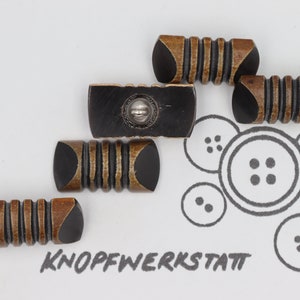 Könnte beinhalten: Fünf braune und schwarze Knöpfe mit einem Metallfuß. Die Knöpfe haben ein gerilltes Design und sind auf einer weißen Oberfläche mit dem Text "KNOFFWERKSTATT" in Schwarz angeordnet.