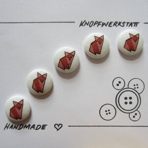 May include: A set of five handmade fabric-covered buttons with a red origami fox design. The buttons are arranged on a white card with the text "KNOPFWERKSTATT" and "HANDMADE". Additional button illustrations are also present.