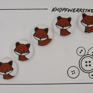 May include: Five brown and white fabric buttons with a fox design. The buttons are arranged in a semi-circle on a white background. The text "KNOFFWERKSTATT" is printed in black at the top of the image.
