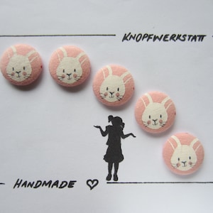 Könnte beinhalten: Sechs rosa Stoffknöpfe mit weißen Hasengesichtern. Die Knöpfe sind in einem Halbkreis auf weißem Hintergrund angeordnet. Der Text "KNOPFWERKSTATT" und "HANDMADE" ist in Schwarz auf dem Hintergrund geschrieben.
