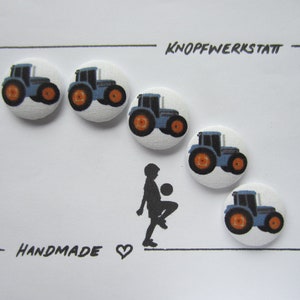 Op de afbeelding: Vijf blauwe en oranje tractor-knoppen met een witte achtergrond. De knoppen zijn in een halve cirkel gerangschikt met de tekst "KNOPFWERKSTATT" en "HANDMADE" eronder.