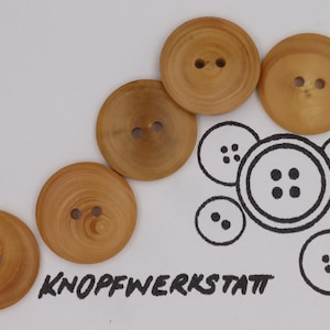 Könnte beinhalten: Fünf Holzknöpfe mit je zwei Löchern, diagonal auf einer weißen Oberfläche angeordnet. Daneben eine Schwarz-Weiß-Illustration von Knöpfen. Der Text "KNOPFWERKSTATT" ist in Schwarz geschrieben.